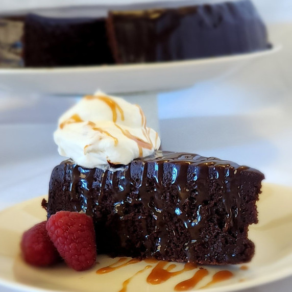 Slice of Deep Dark Rich Chocolate Fudge Cake with caramel drizzle, whipped cream and raspberries
