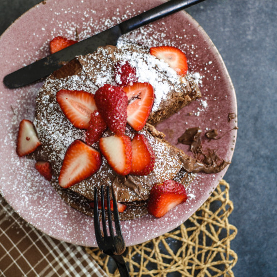 A tall stack of golden pancakes drizzled with Nutella and topped with fresh strawberries.
