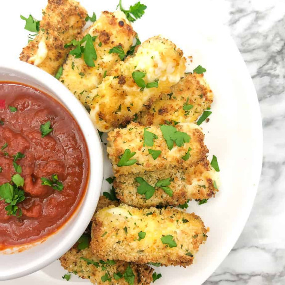 Mozzarella cheese sticks on a plate overhead next to a bowl of sauce topped with parsley