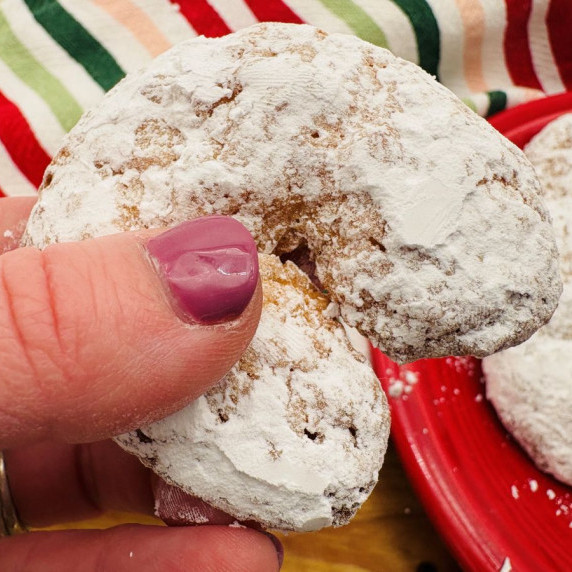 A hand with purple nail polish holds a crescent-shaped cookie covered in powdered sugar.