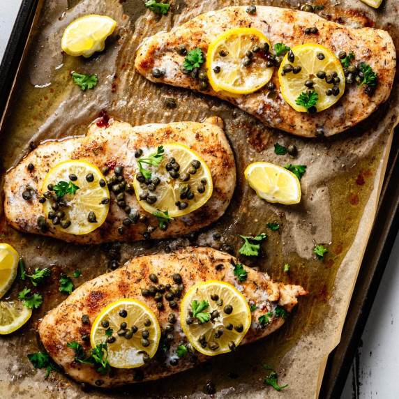 Overhead view of baked chicken piccata on a parchment-lined sheet pan topped with lemon slices, cape