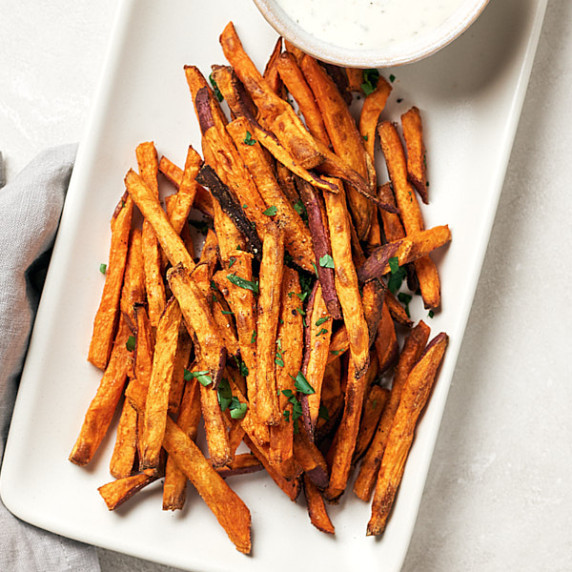 crispy baked sweet potatoes on white platter with small bowl of dip on grey surface