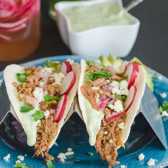 A pair of brisket tacos topped with radish and avocado crema on a blue plate