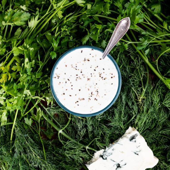 A bowl of  blue cheese dip with a spoon in it, surrounded by fresh herbs. 