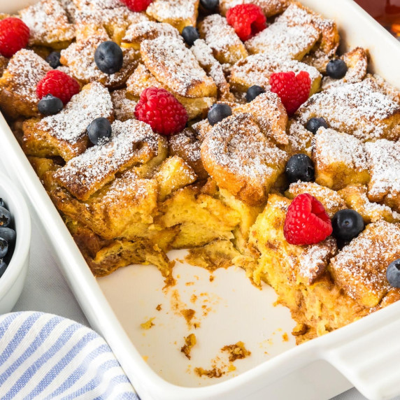 Brioche French toast casserole in a baking pan missing a scoop topped with berries & powdered sugar.