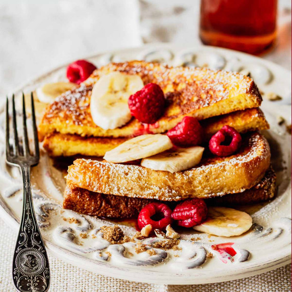 Brioche French toast topped with fresh banana slices and raspberries on a white plate with a fork.