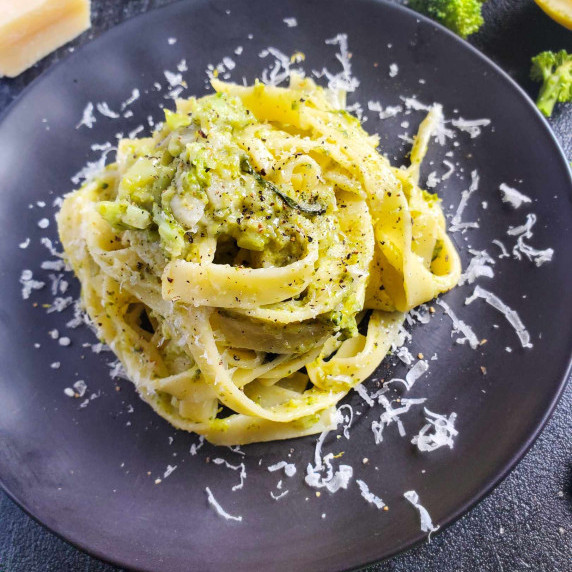A nest of tagliatelle and broccoli on a black plate dressed with white shredded cheese.
