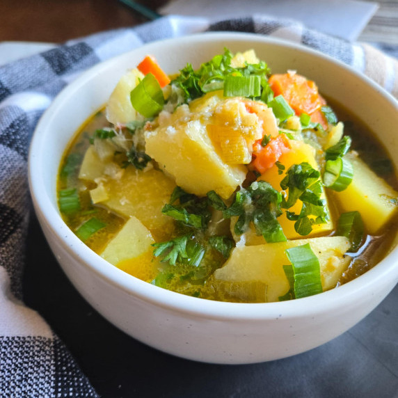 A bowl of potato soup with pops of greens and orange carrots in a white bowl on a black plate