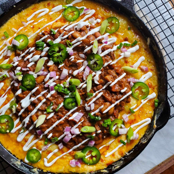 A skillet full of cheesy gnocchi topped with rich chili, white sour cream, greens, and red onions