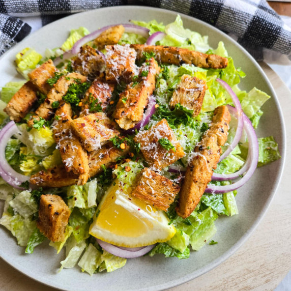 A plate of salad topped off with golden chik'n and croutons, pops of red onion, and a lemon