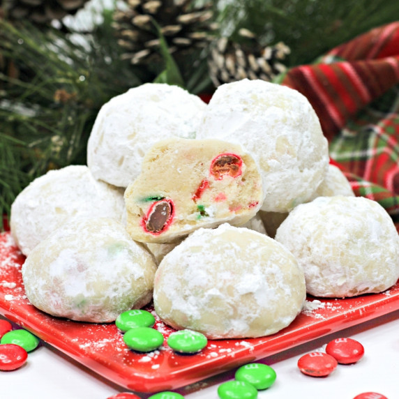 Snowball cookies stacked on a red plate. One of the cookies is cut in half to show M&Ms inside