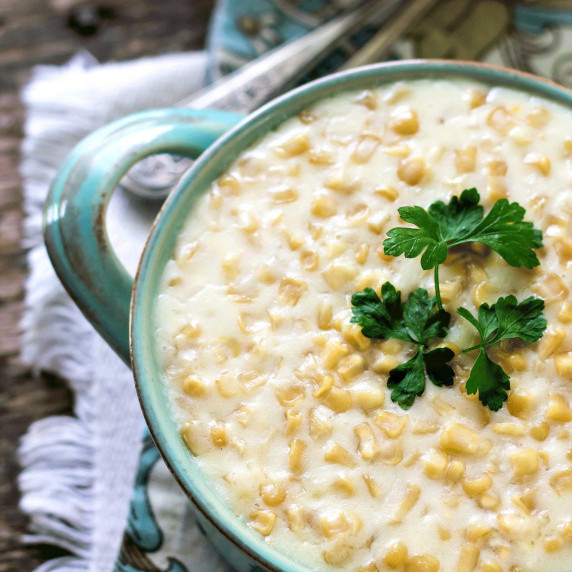 Creamy corn in an antique blue bowl topped with parsley on top of a wooden table