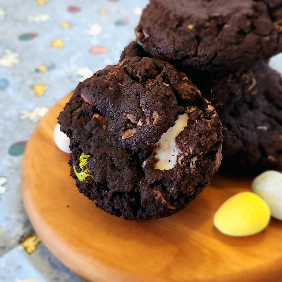 Dark chocolate Easter cookies stacked on a wooden board with Easter eggs and Easter print background