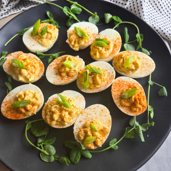 Deviled eggs dusted with red paprika and green onions on a black plate with pea shoot garnishes