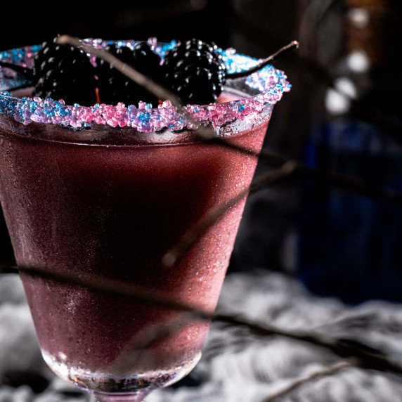 A purple drink in a colorful sugar rimmed glass with blackberries on top.