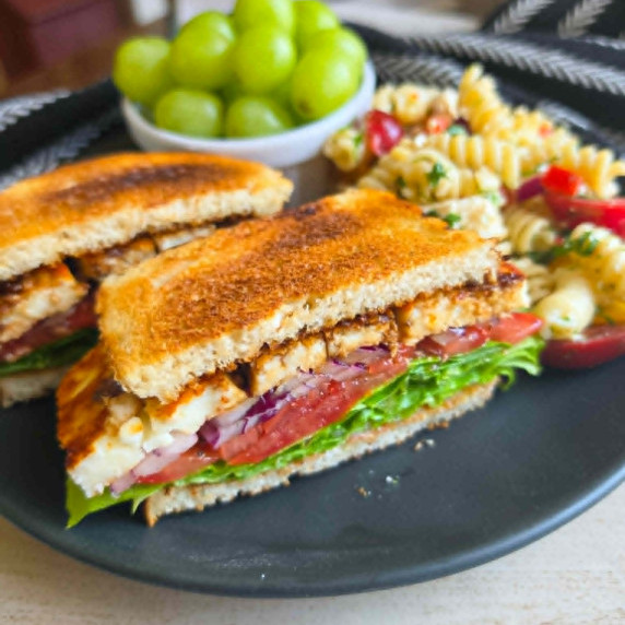 Veggie halloumi sandwich on a black plate with green grapes & pasta salad