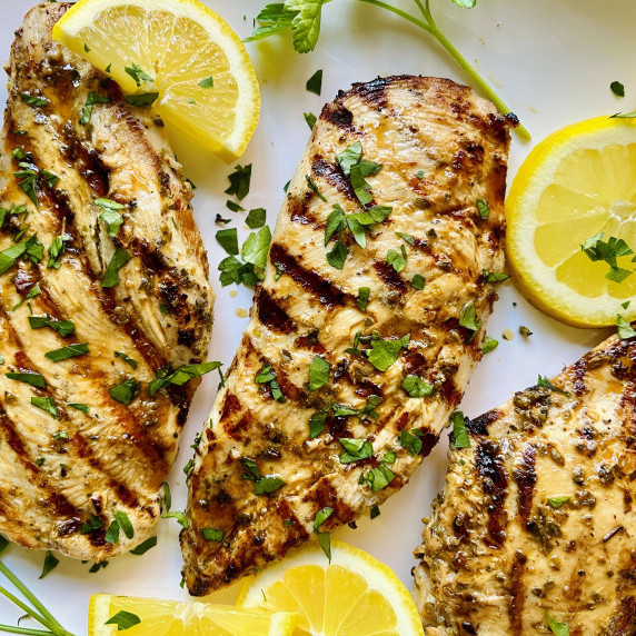Three grilled chicken breasts on a white platter garnished with lemon slices and minced parsley.