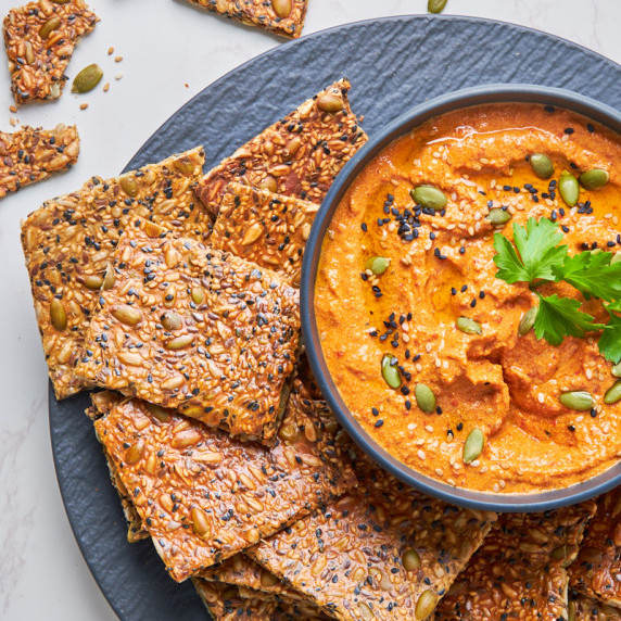 A plate of Seeded Protein Crackers with Sourdough Discard from the top, beside a bright red dip.