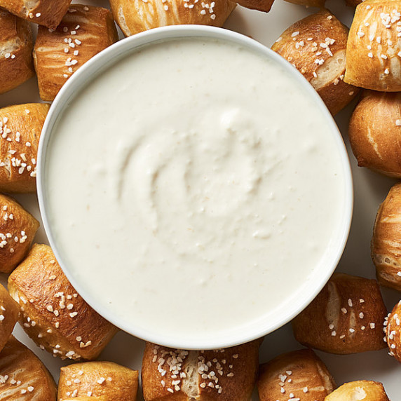 A white bowl with horseradish aioli surrounded by pretzel bites on light background