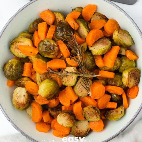 Chopped roasted brussel sprouts and carrots with rosemary on top in a white bowl