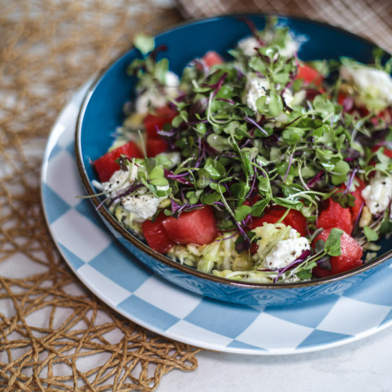 A colorful salad with diced watermelon, shredded cabbage, microgreens, and feta in blue bowl