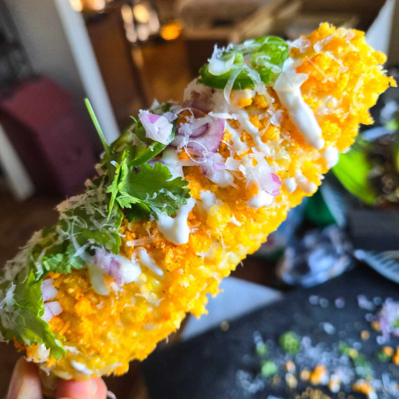 A gorgeous yellow cob of corn with green herbs, white sour cream, and red onions
