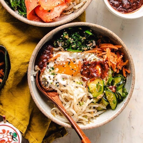 Bean sprouts, spinach, carrots, zucchini, egg, scallions, sesame seeds in a beige bowl with spoon
