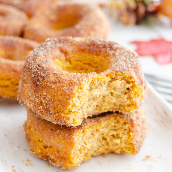 Two keto pumpkin spice donuts with a bite out of them on a white plate. 