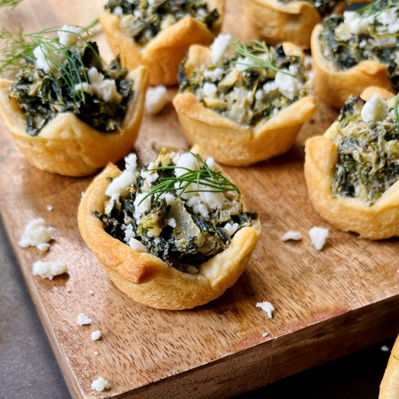 Spinach and cheese tarts topped with feta cheese and fresh dill on a wood cutting board.