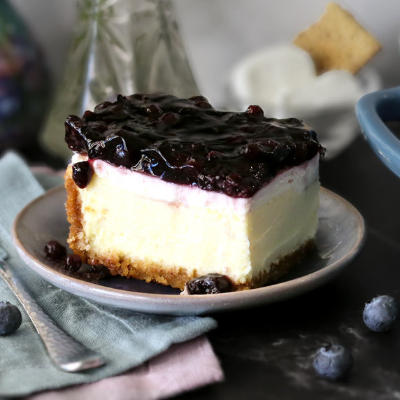 A slice of blueberry topped cheesecake on a small plate with the pan of cheesecake in the background