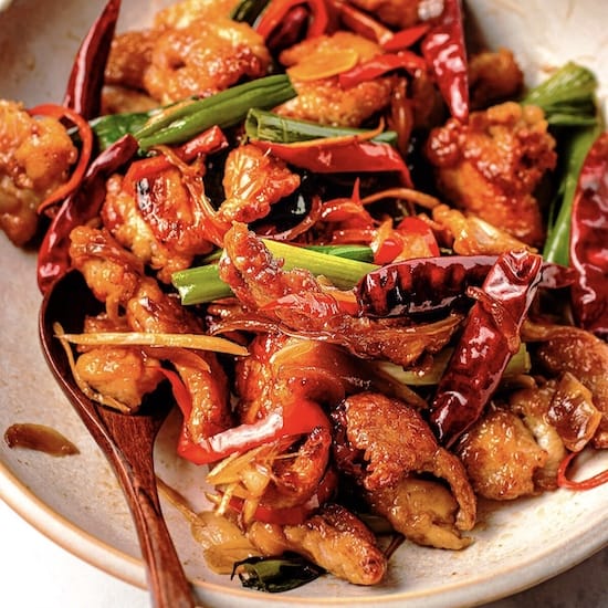 Chicken, red chili peppers and green onions in a white bowl with a wooden spoon