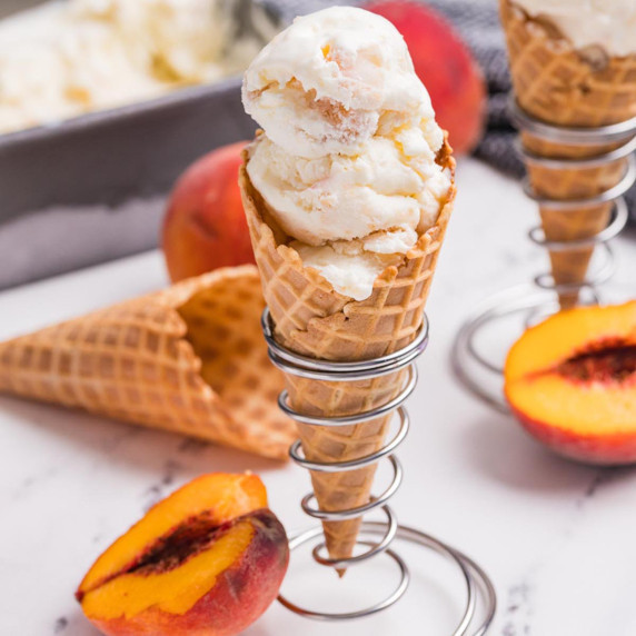 Homemade peach ice cream scooped into waffle cones with fresh peaches and a loaf pan of ice cream in