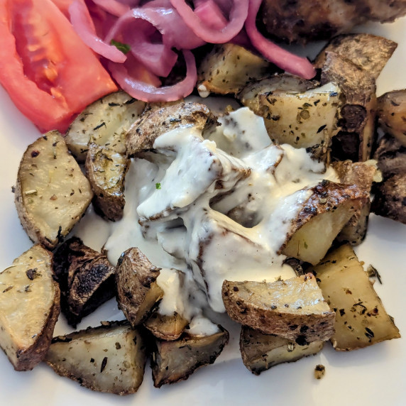 Roasted potatoes with garlic sauce poured over them and tomato slices and pickled onions behind.