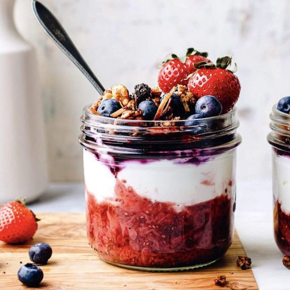 Strawberries, blueberries, granola, yogurt and jam in a jar with a spoon on a wooden cutting board