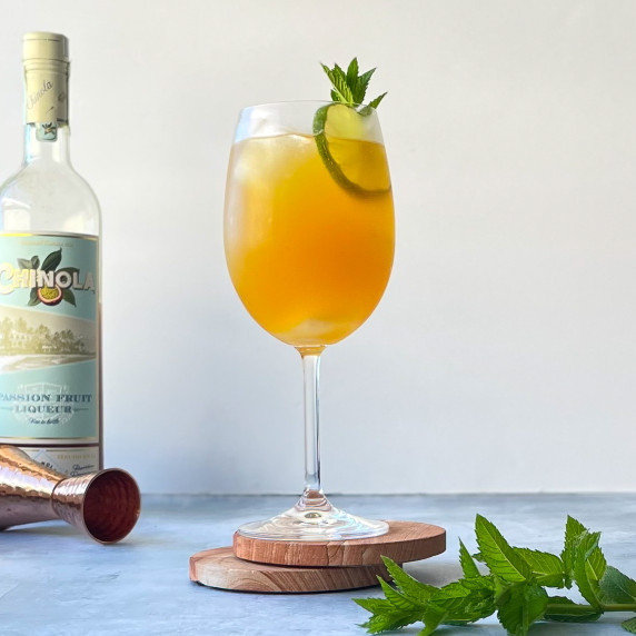 wine glass with passion fruit cocktail garnished with mint and lime next to bottle and mint leaves.