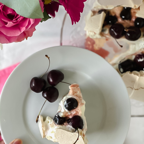 Slice of Pavlova topped with cherry on a white plate.