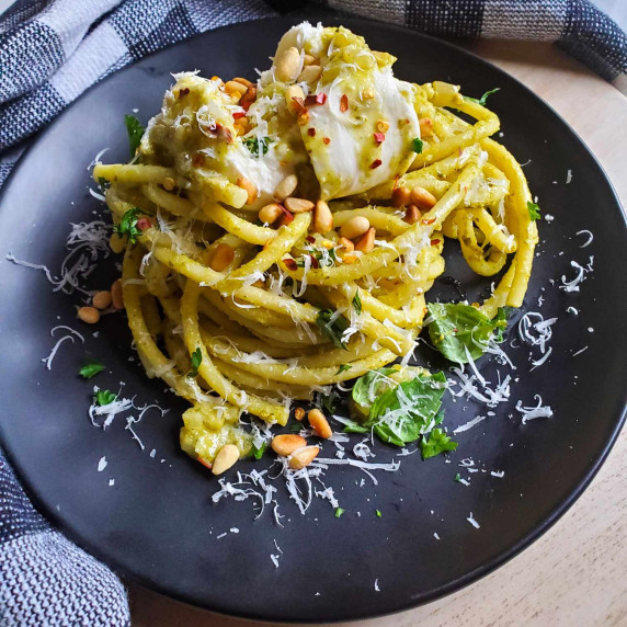 Green pesto bucatini topped with golden brown pine nuts and white burrata cheese on a black plate.
