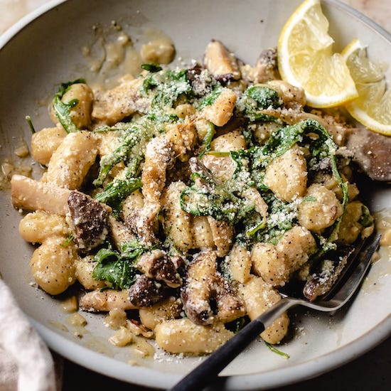 Gnocchi, mushriims, arugula and parmesan cheese on a white plate with a fork and lemon slices