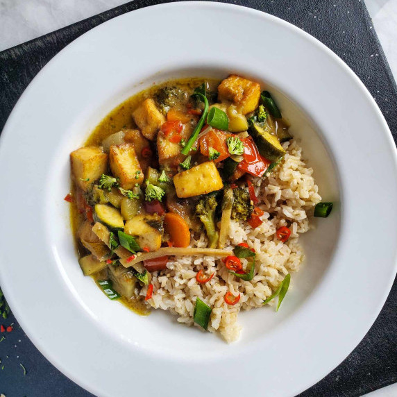 Green curry and brown rice in a white rimmed bowl on a black cutting board.
