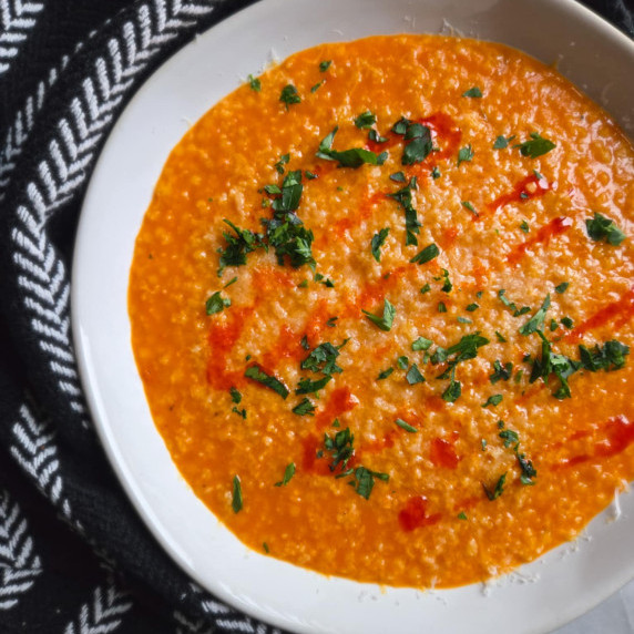 A bright red pepper & noodle soup with green parsley and chili oil against a black towel.