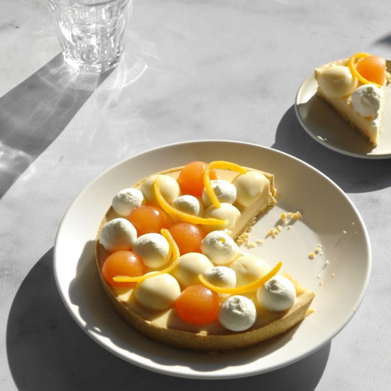 A grapefruit tart with a slice cut out of it sits on a white marble backdrop with hard shadows.
