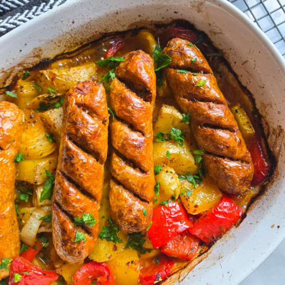 Scored sausages on a bed of colourful veggies in a white baking dish.