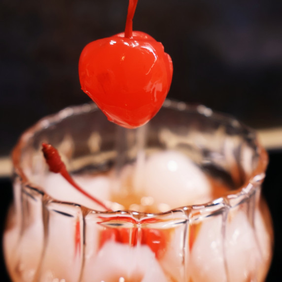 A maraschino cherry is held above a glass filled with ice and Shirley Temple cocktail.