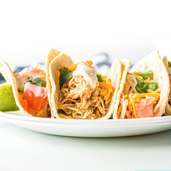 four tacos on a plate loaded with shredded chicken, cheese, sour cream and cilantro