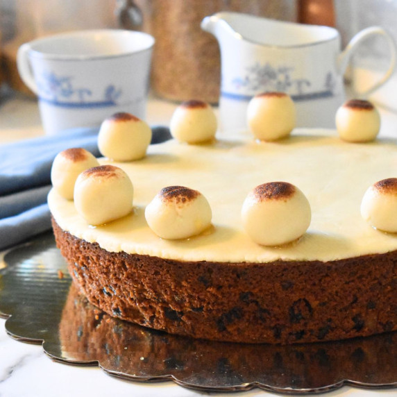 A simnel cake, topped with eleven marzipan balls, with some tea cups in the background