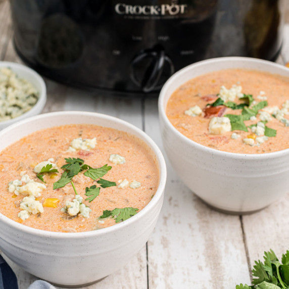 two bowls of buffalo chicken chili in front of a slow cooker with blue cheese and cilantro sprinkled
