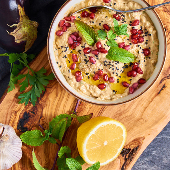 A bowl filled with Smoked Aubergine Dip on a wooden board. Topped with Pomegranate seeds and sesame.