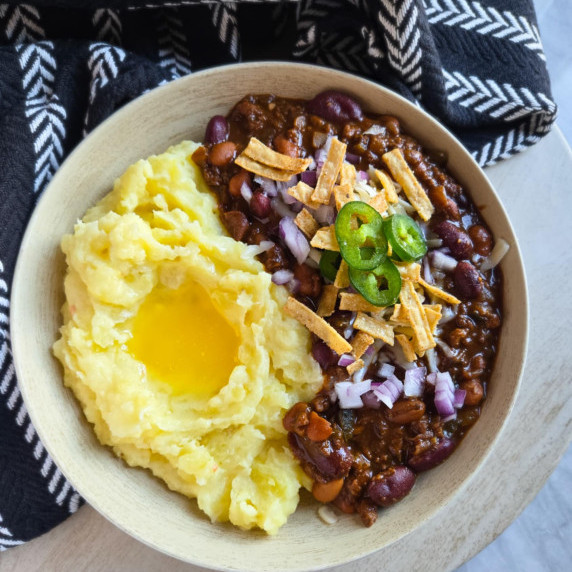 A bowl of creamy mashed potatoes and rich, vibrant chili against a black towel.