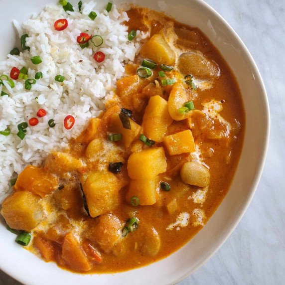 A white bowl full of potato and bean red curry with green onions and red chilis.