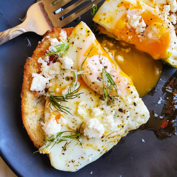 Fried egg on sourdough toast with green dill & crumbled feta against a black plate.